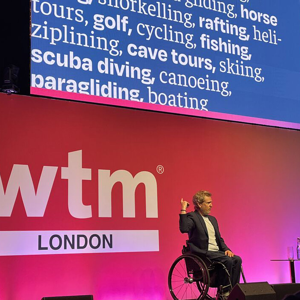 Ryan on stage in London pointing to a slide behind him listing many accessible outdoor activities